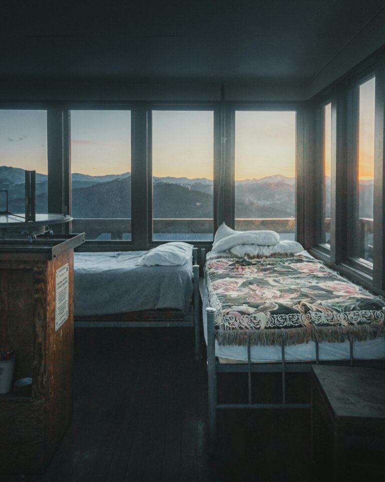 Cozy mountain cabin bedroom with panoramic window view of sunset over peaks. Rustic interior blends comfort with nature.